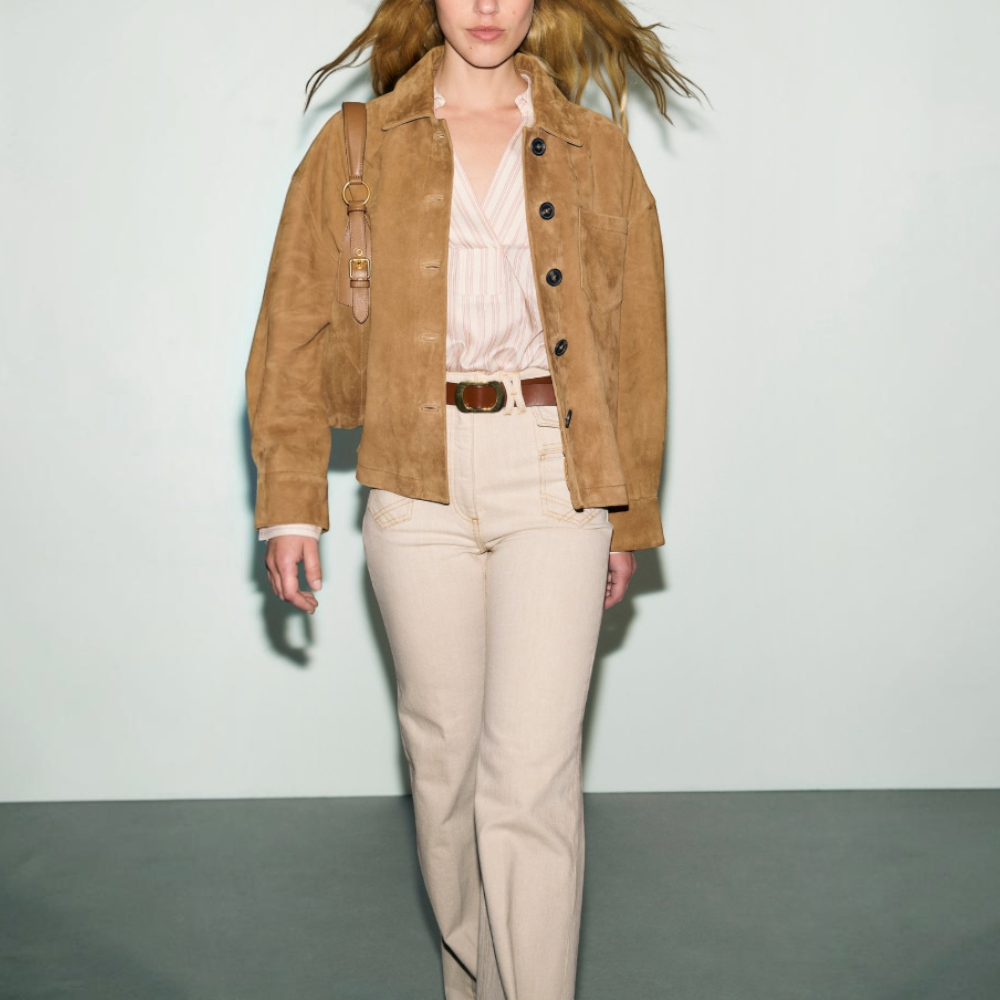 A person with long blonde hair wears a veste en cuir velours camel, a striped shirt, beige high-waisted pants, and a brown belt, carrying a tan shoulder bag while walking against a light background.